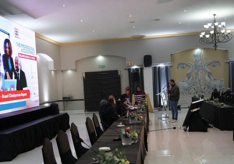 Large LED screen showing a presentation beside a boardroom-style table layout with a camera operator at the lectern
