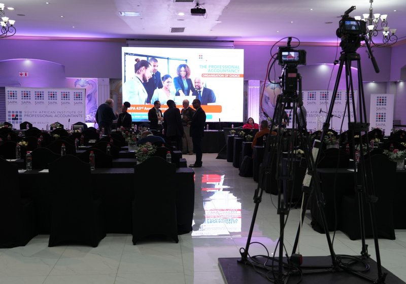 Conference hall with rows of tables, a large LED screen, brand banners and two video cameras on tripods in the foreground
