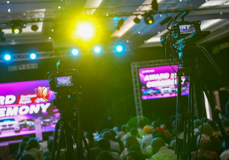 Two video cameras on tripods filming a live awards event with a large LED screen behind the stage