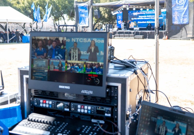 Flight case rack with two recorders, a switcher and a multiview monitor at an outdoor event, with the stage and seated audience in the background