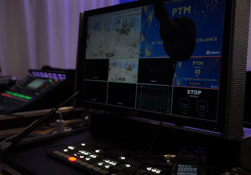 Vision mixer control surface below a multiview monitor showing camera feeds, a presentation slide and recorder status, with headphones hanging on the screen