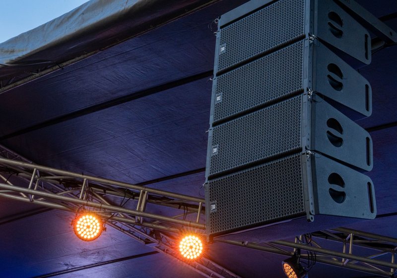 Line-array speaker cabinets and glowing LED par lights hung from truss under an outdoor stage roof