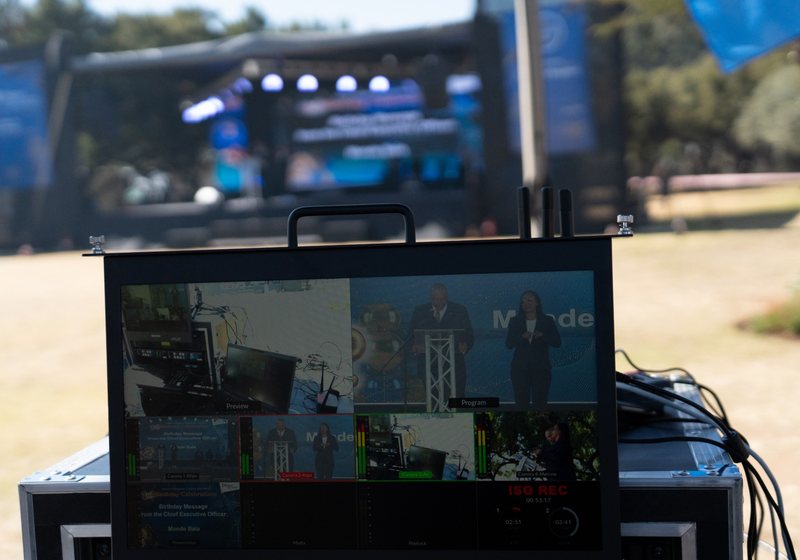 Multiview vision mixer monitor showing four camera feeds on a flight case, with an outdoor stage blurred behind