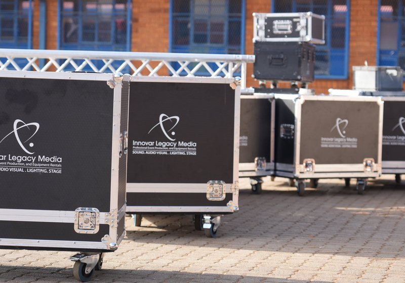 Row of Innovar Legacy Media branded flight cases on castors on brick paving with a truss section behind