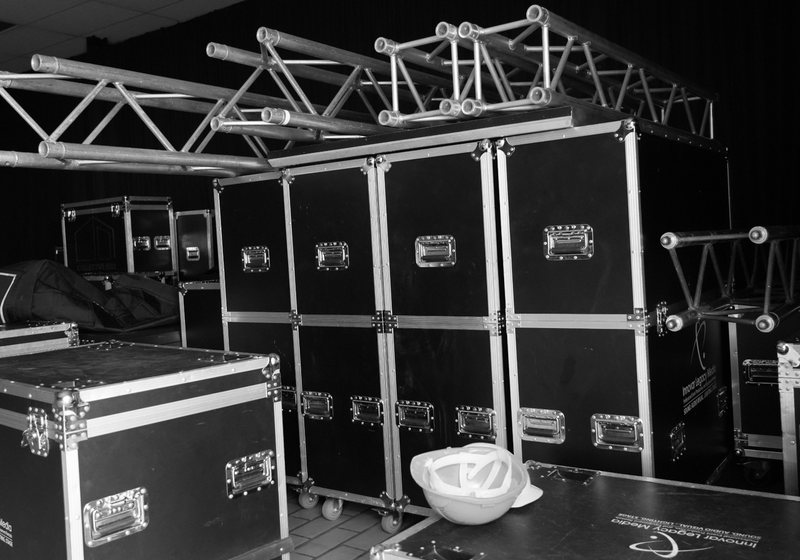 Black and silver flight cases stacked in a store room with truss sections resting on top, in black and white