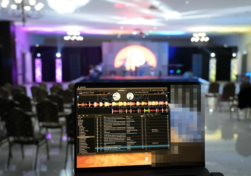 DJ software on a laptop in the foreground with a stage, lit truss columns and rows of chairs blurred behind
