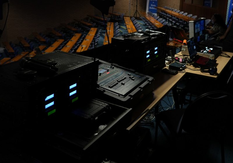 Front of house position at the back of a tiered auditorium with wireless microphone racks, a mixing console and a laptop desk, with a welcome slide on the screen