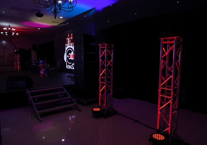 Stacked line-array speakers beside two red-lit truss towers with LED par lights, a stage and a screen in a dim venue