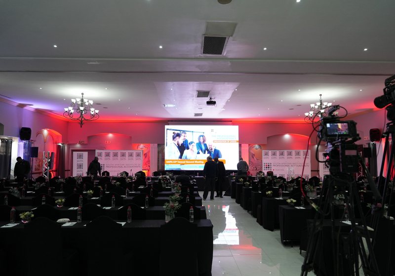 Conference hall with rows of black-draped tables, a large LED screen and branded backdrops, with video cameras on tripods in the foreground