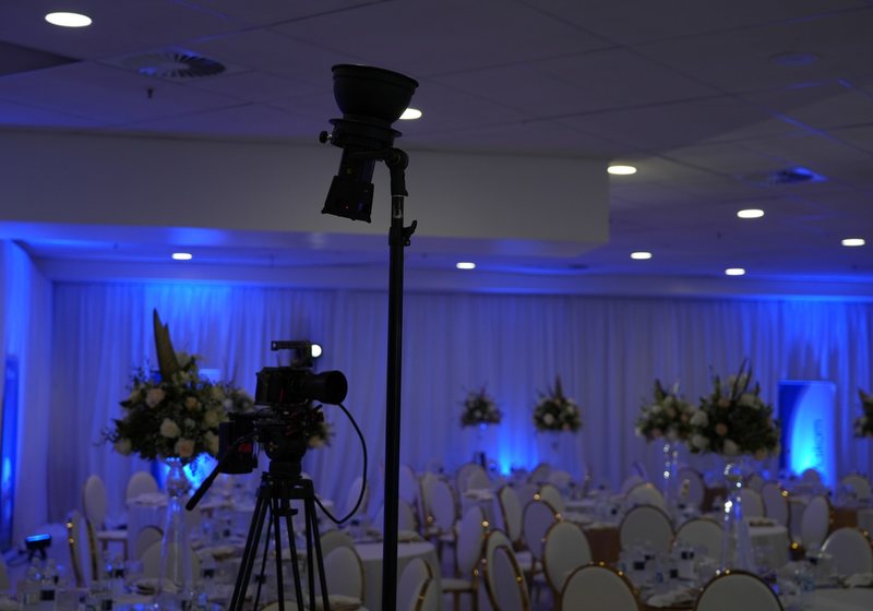 Banquet hall with round tables, tall floral centrepieces and blue uplit drapes, with a video camera on a tripod and a light on a stand in the foreground