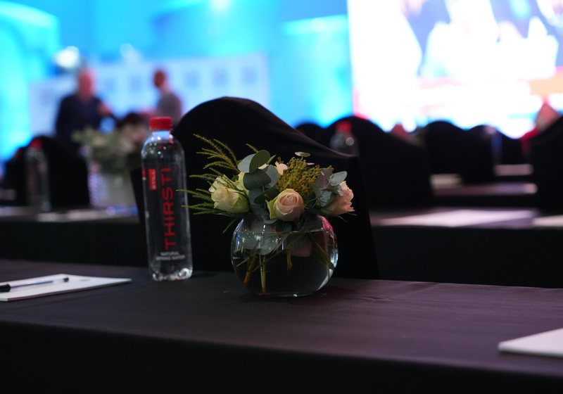 Vase of roses and eucalyptus and a water bottle on a black-draped conference table with a bright screen blurred behind