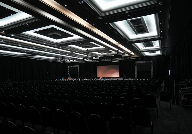 Large dark conference hall with lit ceiling panels, hundreds of chairs facing a stage with truss frames, screens and lighting