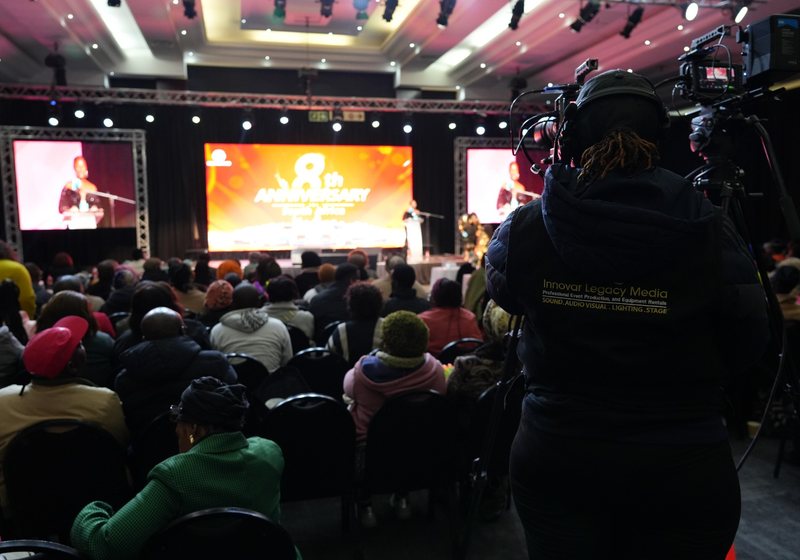 Innovar camera operator behind two cameras among a seated audience facing a stage with a large LED screen and two side screens