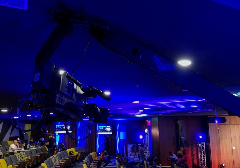 Broadcast camera on a ceiling-height jib arm in a blue-lit auditorium with a control table, monitors and truss lights