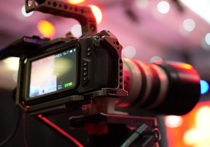 Close-up of a cinema camera in a metal cage with a flip-out monitor and long lens against pink event lighting