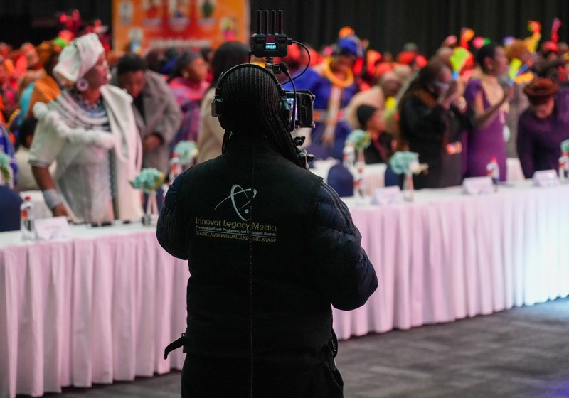 Innovar camera operator with headphones and a wireless video transmitter on the camera, facing long banquet tables and guests in colourful dress
