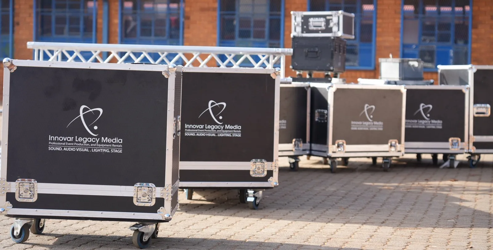 A row of Innovar Legacy Media branded flight cases on castors on brick paving with a truss section behind