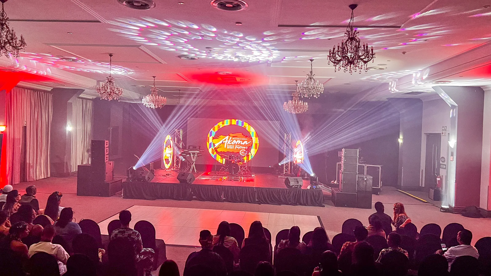 An audience seated in a ballroom facing a stage with a round LED screen and two tall side LED screens, lit by beam lights