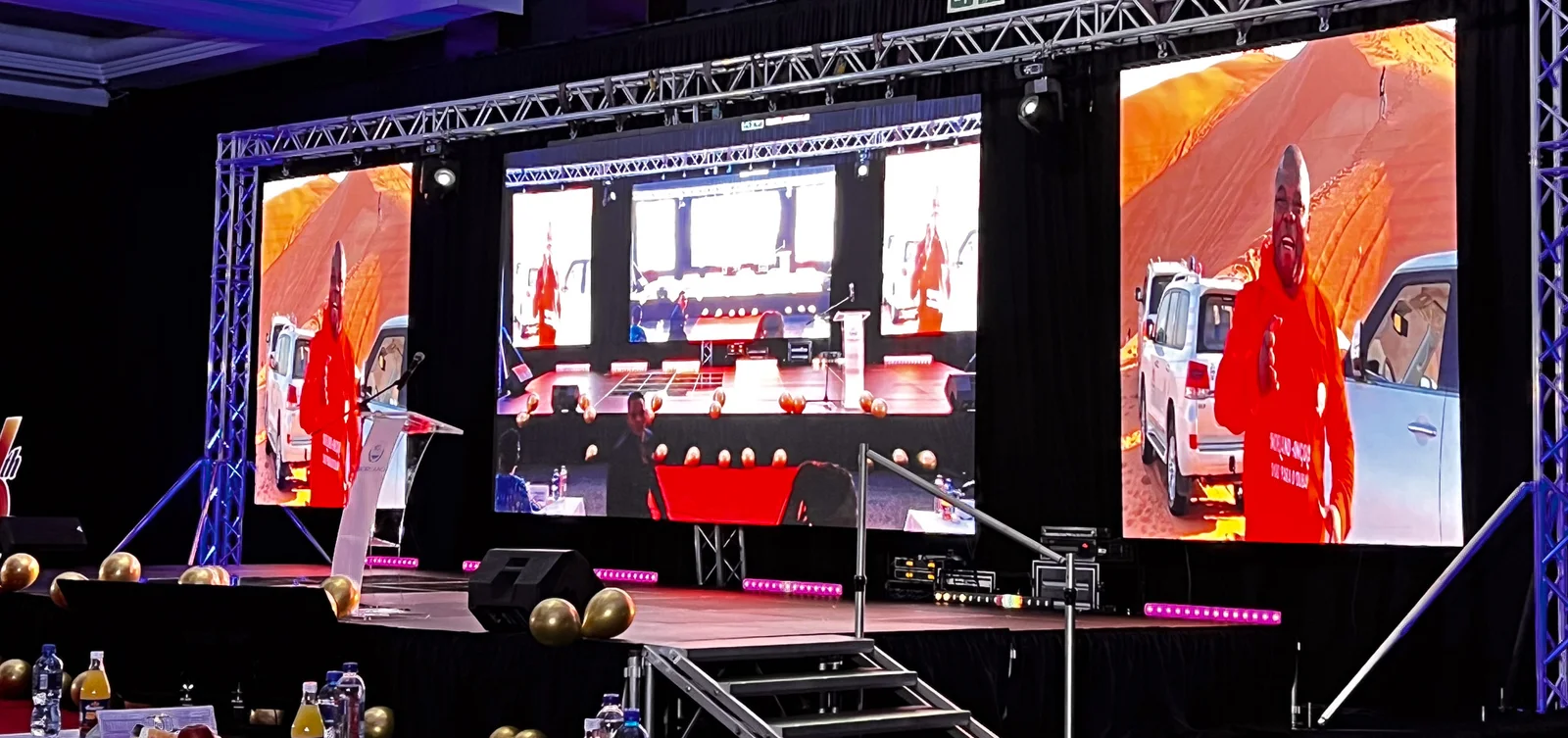 A stage with a large centre LED screen and two tall side LED screens on a truss, showing a presenter on the side screens and a wide view of the room on the centre screen